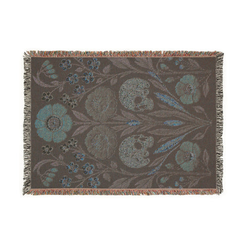 Woven cotton throw blanket with fringed edges and a black ground. Two cream skulls sit side by side at the center of a symmetrical folk garden. A terracotta poppy crowns the top, plum peonies bloom in the middle, and clusters of golden-yellow and white wildflowers, green leaves, and wheat-like sprigs fill the field, with more terracotta poppies along the bottom. The dimensional pattern is woven through a jacquard process, and the reverse shows a mirrored, softer version of the design.