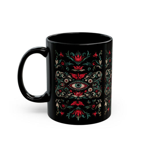 A black ceramic mug featuring a symmetrical folk art design with a central mystic eye motif in turquoise and coral. The design includes ornate scrollwork, red tulip-like flowers, sage green leaves, and cream flourishes arranged in a decorative pattern. The mug has a glossy finish with a C-shaped handle.