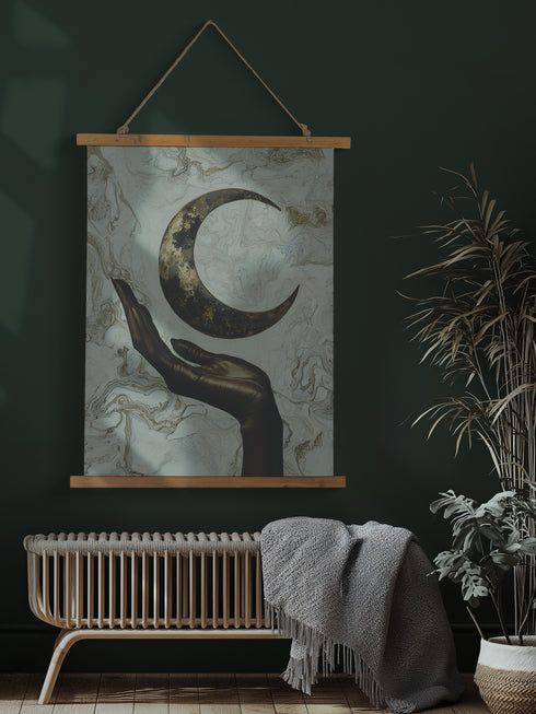 A wall tapestry featuring a bronze-colored hand reaching upward toward an antiqued gold crescent moon, set against a swirling ivory and gold marbled background, mounted between wooden dowels with twine.