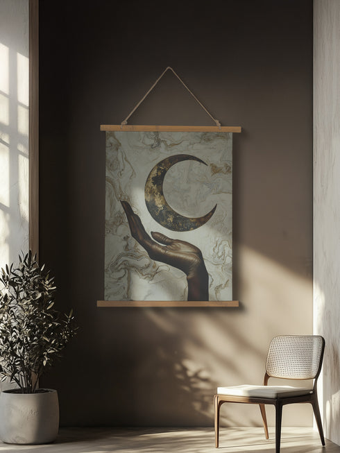 A wall tapestry featuring a bronze-colored hand reaching upward toward an antiqued gold crescent moon, set against a swirling ivory and gold marbled background, mounted between wooden dowels with twine.