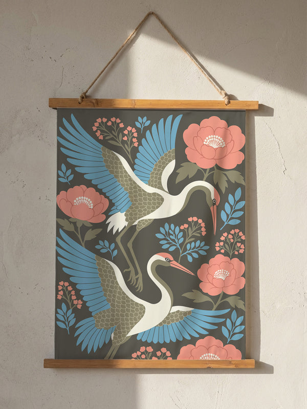 Vintage style Chinoiserie crane wall hanging by Grimoire and Goblet featuring two white cranes with light blue wings flying among coral pink peonies and sage green leaves against a dark charcoal grey background. The smooth polyester tapestry is suspended from artisanal wooden dowels with a natural twine hanger. A Grimoire and Goblet exclusive botanical design perfect for adding moody, Dark Academia ambiance to any apartment or sanctuary.