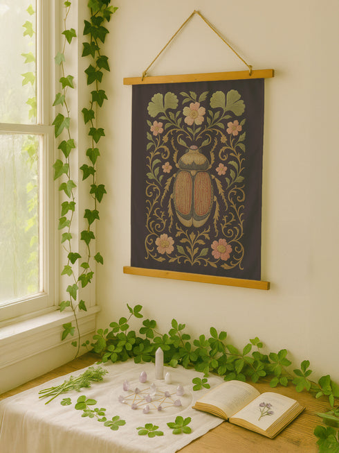 26x36 inch vertical wall tapestry printed on silky polyester fabric, featuring a detailed embroidered-style beetle surrounded by pink and green florals and curling botanical vines on a black background, with wooden hanger and twine at top and bottom. Art nouveau, vintage-inspired, cottagecore, gothic decor.