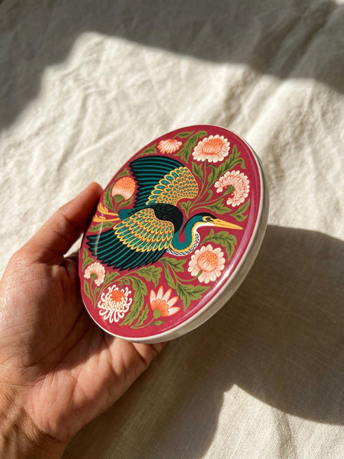 A round, 4-inch ceramic drink coaster featuring a glossy, flat-printed Art Nouveau design. The central motif is a flying heron with teal plumage and scalloped golden wing feathers, surrounded by stylized winding olive green foliage, peach chrysanthemums, and botanical blooms. The artwork is set against a solid, deep blood-red background. The coaster has a smooth white edge and a natural brown cork backing. Available as a single coaster or in matching sets of four or six.
