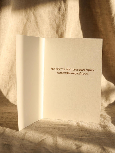 A vintage-style greeting card titled "DE HUMANI CORDIS" featuring a medical illustration of two anatomical hearts connected by a single looped blood vessel. The hearts are drawn in a sepia and crimson woodcut style on a cream background mimicking aged parchment. Text below reads "Fig. 14: The Twin Pulse." The card evokes a dark academia or cottagecore aesthetic. Inside text reads "Two different beats, one shared rhythm.