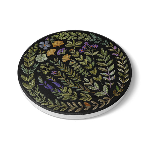 Circular ceramic coaster with black background featuring intricate botanical illustrations of various herbs, flowers and leaves in green, purple and gold colors with white ceramic edge and cork backing.