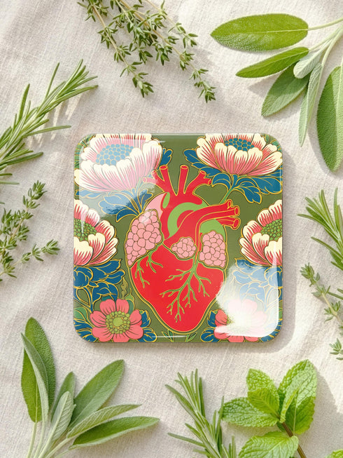 A set of four premium 4x4 inch anatomical heart coasters from Grimoire and Goblet. Each square coaster is made of masonite hardboard with a high-gloss finish and a non-slip cork base. The designs feature intricate biological heart illustrations with folk-art floral motifs in vibrant crimson, pink, yellow, and purple against mossy green and deep teal backgrounds. The high-fidelity print mimics a luxury embroidered texture, perfect for a cardiology gift or dark academia home decor.