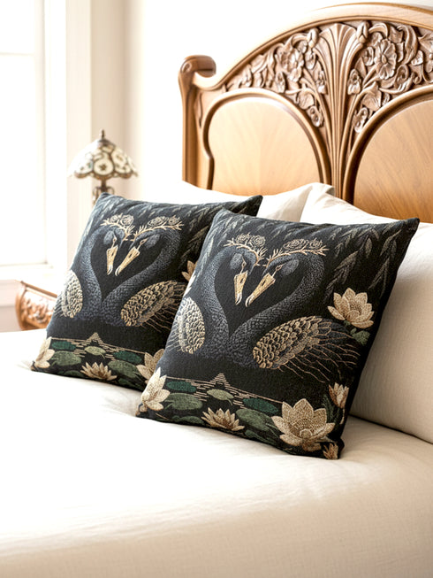 A double-sided woven jacquard throw pillow by Grimoire and Goblet measuring 17 by 18 inches. The tactile tapestry design features two black swans facing each other, their gracefully curved necks forming a subtle heart shape. Each swan wears a delicate floral crown in gold and cream tones. They float on a dark pond adorned with muted green lily pads and cream water lilies. The textured surface is woven from cotton and polyester threads.