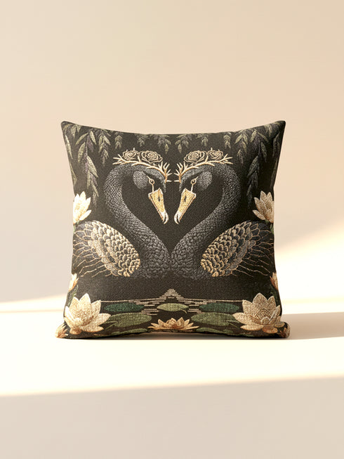 A double-sided woven jacquard throw pillow by Grimoire and Goblet measuring 17 by 18 inches. The tactile tapestry design features two black swans facing each other, their gracefully curved necks forming a subtle heart shape. Each swan wears a delicate floral crown in gold and cream tones. They float on a dark pond adorned with muted green lily pads and cream water lilies. The textured surface is woven from cotton and polyester threads.