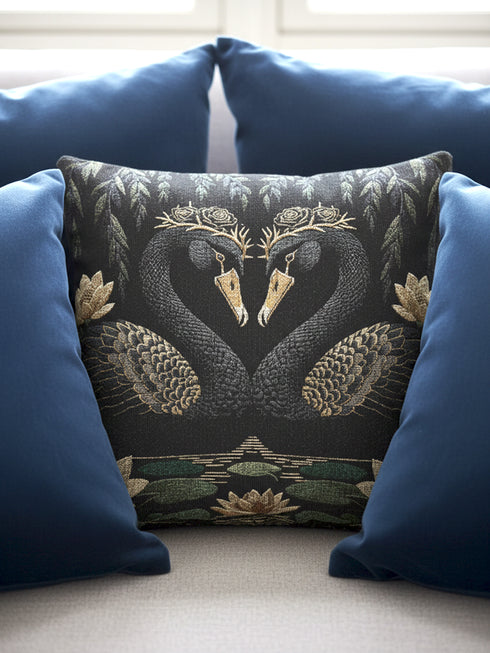 A double-sided woven jacquard throw pillow by Grimoire and Goblet measuring 17 by 18 inches. The tactile tapestry design features two black swans facing each other, their gracefully curved necks forming a subtle heart shape. Each swan wears a delicate floral crown in gold and cream tones. They float on a dark pond adorned with muted green lily pads and cream water lilies. The textured surface is woven from cotton and polyester threads.