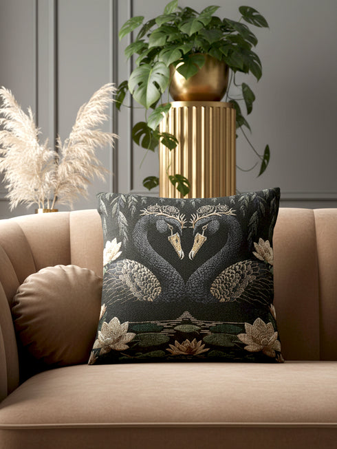 A double-sided woven jacquard throw pillow by Grimoire and Goblet measuring 17 by 18 inches. The tactile tapestry design features two black swans facing each other, their gracefully curved necks forming a subtle heart shape. Each swan wears a delicate floral crown in gold and cream tones. They float on a dark pond adorned with muted green lily pads and cream water lilies. The textured surface is woven from cotton and polyester threads.