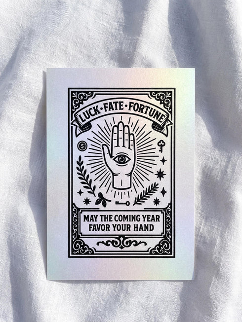Grimoire and Goblet presents a 5x7 inch pearlescent greeting card. The design features a black linocut-style illustration of an open palm with an eye, surrounded by rays, keys, coins, and stars. The cover reads Luck Fate Fortune and May The Coming Year Favor Your Hand. The interior features a smaller hand icon and a printed blessing. The paper has a luxury silvery shimmer finish.
