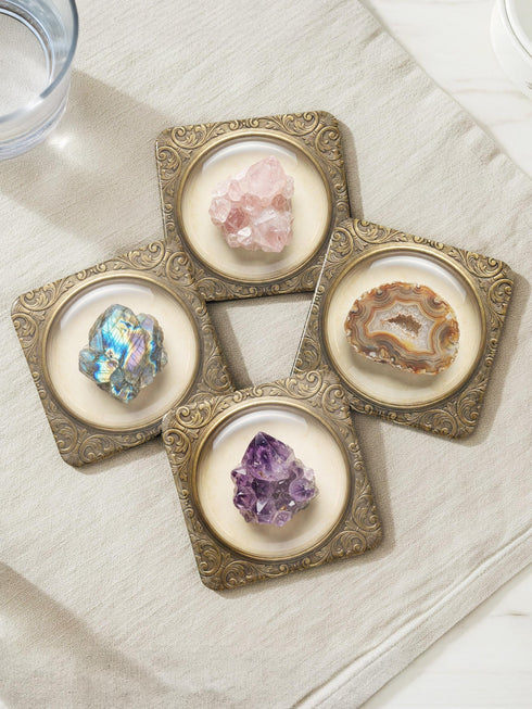 Grimoire and Goblet set of four square glossy coasters featuring high-resolution printed illustrations of raw crystals: purple amethyst, pink rose quartz, iridescent labradorite, and banded agate. Each gemstone is depicted inside a printed faux antique brass baroque frame with intricate scrollwork, set against a cream parchment background. The design is a flat 2D trompe l'œil print on hardboard, creating a realistic 3D specimen box illusion.