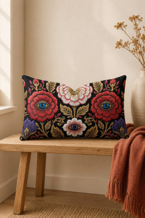 Evil eye lumbar pillow cover in black featuring a symmetrical folk art floral print. Two crimson red roses hold bright blue eyes at their centers, a white blossom below cradles a violet purple eye, and antique gold leaves, scrollwork, and pink buds frame the design. Purple tulips anchor the lower corners. Rectangular 20 x 12 inch cushion case with a hidden zipper and the double sided embroidered look design printed on smooth, soft, linen feel polyester fabric.