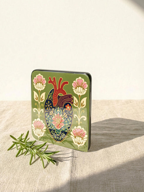 A set of four premium 4x4 inch anatomical heart coasters from Grimoire and Goblet. Each square coaster is made of masonite hardboard with a high-gloss finish and a non-slip cork base. The designs feature intricate biological heart illustrations with folk-art floral motifs in vibrant crimson, pink, yellow, and purple against mossy green and deep teal backgrounds. The high-fidelity print mimics a luxury embroidered texture, perfect for a cardiology gift or dark academia home decor.