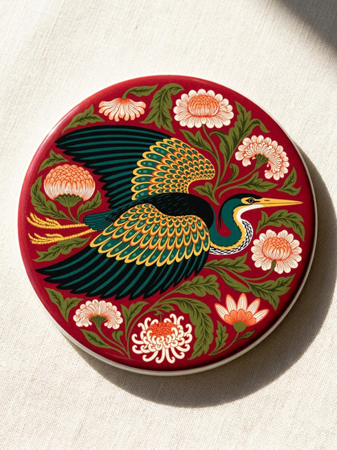 A round, 4-inch ceramic drink coaster featuring a glossy, flat-printed Art Nouveau design. The central motif is a flying heron with teal plumage and scalloped golden wing feathers, surrounded by stylized winding olive green foliage, peach chrysanthemums, and botanical blooms. The artwork is set against a solid, deep blood-red background. The coaster has a smooth white edge and a natural brown cork backing. Available as a single coaster or in matching sets of four or six.