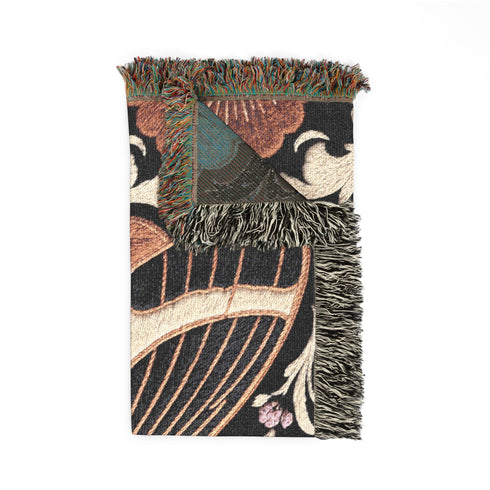 Woven cotton throw blanket with fringed edges and a black ground. At the center is a large moth with copper, brown, and cream wings marked by fine stripes and a concentric eyespot, with feathered antennae. Cream acanthus scrollwork fans out around it in the Art Nouveau manner, joined by clusters of lilac and mauve flowers and copper-brown blossoms with pale rays at the top and bottom. The pattern is jacquard woven, and the reverse shows a mirrored, softer version of the design.