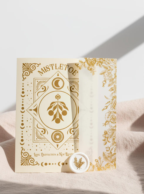 A Grimoire & Goblet luxury Mistletoe ritual card set. The 5x7 cream greeting card features gold celestial designs framing “Mistletoe” and “Love, Protection & New Beginnings.” Shown with a vellum wrap printed in gold florals, a white wax seal with mistletoe, and a premium envelope. Includes a 4x6 ritual insert on handmade deckled paper titled “Ritual for Peace and Protection.” A spiritual Yule stationery set designed as a luxurious unboxing experience.