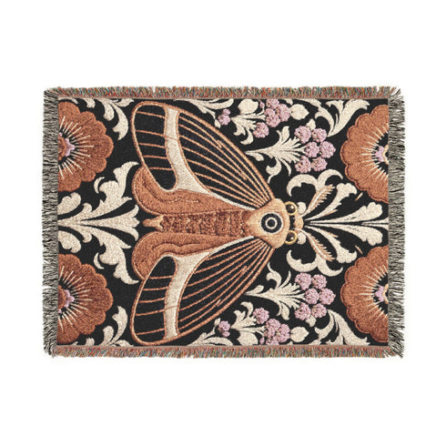 Woven cotton throw blanket with fringed edges and a black ground. At the center is a large moth with copper, brown, and cream wings marked by fine stripes and a concentric eyespot, with feathered antennae. Cream acanthus scrollwork fans out around it in the Art Nouveau manner, joined by clusters of lilac and mauve flowers and copper-brown blossoms with pale rays at the top and bottom. The pattern is jacquard woven, and the reverse shows a mirrored, softer version of the design.