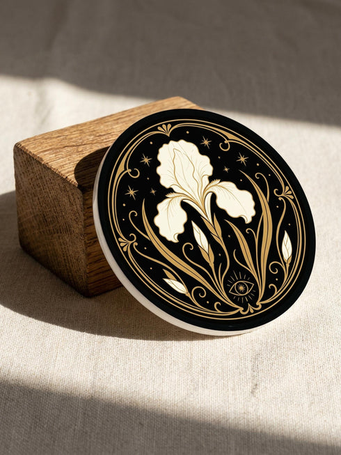 A set of four round 4-inch ceramic coasters featuring a glossy 2D flat-printed Art Deco botanical design. Each coaster displays a different cream-colored flower including a moonflower, foxglove, poppy, and iris, set against a solid black background. The floral motifs are framed by intricate gold linework, crescent moons, four-pointed stars, and mystical symbols like a candle and an all-seeing eye. The coasters have a smooth white edge and are finished with a natural brown cork backing.