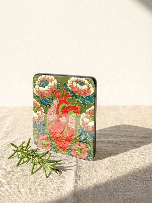 A set of four premium 4x4 inch anatomical heart coasters from Grimoire and Goblet. Each square coaster is made of masonite hardboard with a high-gloss finish and a non-slip cork base. The designs feature intricate biological heart illustrations with folk-art floral motifs in vibrant crimson, pink, yellow, and purple against mossy green and deep teal backgrounds. The high-fidelity print mimics a luxury embroidered texture, perfect for a cardiology gift or dark academia home decor.