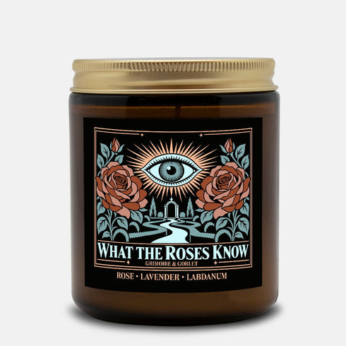 A 9 oz amber glass jar candle with a brushed gold screw lid and a wraparound black label. The label shows an all-seeing eye radiating peach and gold rays above a winding garden path that leads through slender cypress trees to an arched gate, flanked by two dusty coral roses on sage green stems, inside a fine copper border. Cream serif text reads What the Roses Know, Grimoire & Goblet, and Rose, Lavender, Labdanum. A romantic, witchy, art nouveau garden design.