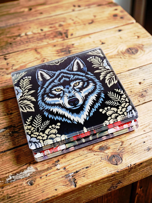 Set of 4 square tempered glass coasters featuring a mystical wolf design on a black background. High-gloss finish with non-slip rubber feet. Dark Academia and Whimsigoth aesthetic home decor. Close-up detail shows grey wolf fur texture and amber eyes. Durable drinkware protection for wooden tables. Grimoire and Goblet gothic luxury barware accessory for coffee tables and desks. Heat resistant glass surface with polished edges.