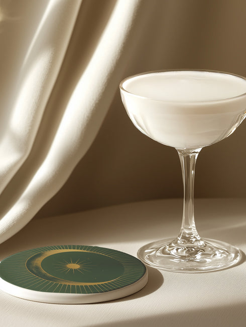 Round ceramic coaster with forest green background. Golden crescent moon embraces a sun at the center. Fine golden lines radiate outward, creating a sunburst pattern to the edge. White rim indicates cork backing. Coaster diameter is 4.04 inches.