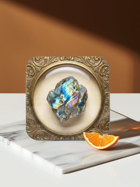 Grimoire and Goblet set of four square glossy coasters featuring high-resolution printed illustrations of raw crystals: purple amethyst, pink rose quartz, iridescent labradorite, and banded agate. Each gemstone is depicted inside a printed faux antique brass baroque frame with intricate scrollwork, set against a cream parchment background. The design is a flat 2D trompe l'œil print on hardboard, creating a realistic 3D specimen box illusion.