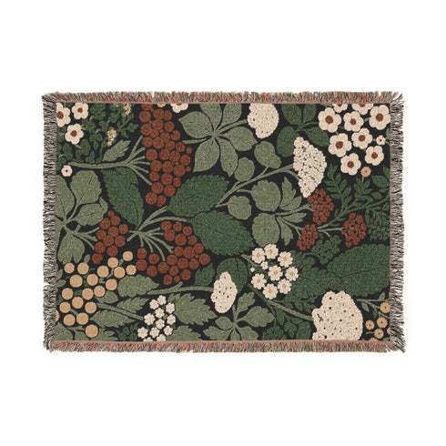 A Grimoire and Goblet luxury woven throw blanket featuring a dark botanical design. The Jacquard cotton tapestry showcases cream elderflower clusters and rust-colored berries against a deep black background. Sage green leaves interweave throughout the floral pattern. The blanket is finished with multicolored fringe edges. A moody cottagecore aesthetic piece ideal for dark academia decor.