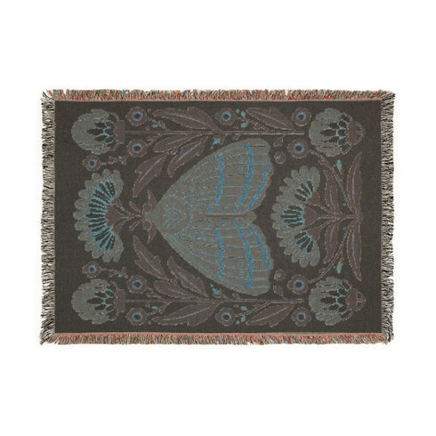 Grimoire and Goblet woven jacquard cotton blanket / wall tapestry featuring a centered folk-art moth framed by symmetrical wildflowers and leafy stems. Natural oatmeal-beige base with rust/terracotta and cocoa-brown moth detail, charcoal-black botanicals, and warm ivory highlights. Finished with a 2-inch multicolor fringe and a reversible color-negative back. Available in 52x37, 50x60, and 60x80 inches as a throw, bed cover, or statement wall hanging.