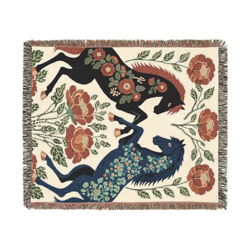 A woven cotton throw blanket by Grimoire and Goblet featuring two rearing horses facing each other in a symmetrical folk art design. One horse is charcoal black with rust floral detailing; the other is navy blue with teal floral detailing. They are framed by large rust-colored peonies and green leafy vines on a cream background. The blanket features a multicolor fringe edge. Perfect for dark academia and cottagecore aesthetics.