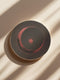 A round ceramic plate with a sleek design featuring a crescent moon and radiating lines in a muted red and black color scheme. The plate is set against a warm brown background with soft lighting, highlighting the celestial and minimalist aesthetic of the piece.
