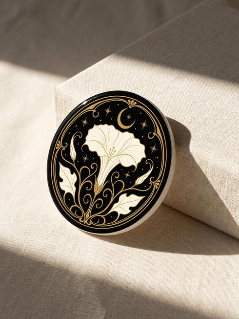 A set of four round 4-inch ceramic coasters featuring a glossy 2D flat-printed Art Deco botanical design. Each coaster displays a different cream-colored flower including a moonflower, foxglove, poppy, and iris, set against a solid black background. The floral motifs are framed by intricate gold linework, crescent moons, four-pointed stars, and mystical symbols like a candle and an all-seeing eye. The coasters have a smooth white edge and are finished with a natural brown cork backing.