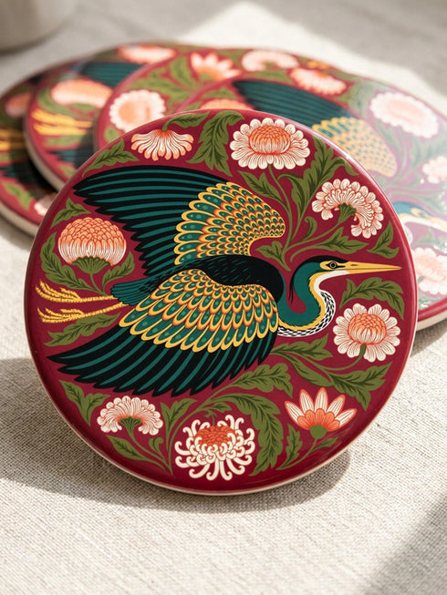 A round, 4-inch ceramic drink coaster featuring a glossy, flat-printed Art Nouveau design. The central motif is a flying heron with teal plumage and scalloped golden wing feathers, surrounded by stylized winding olive green foliage, peach chrysanthemums, and botanical blooms. The artwork is set against a solid, deep blood-red background. The coaster has a smooth white edge and a natural brown cork backing. Available as a single coaster or in matching sets of four or six.