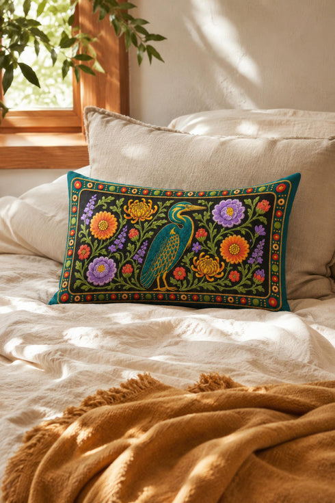 Heron lumbar pillow cover featuring a teal wading bird with gold detailing standing among orange marigolds, golden chrysanthemums, purple peonies, lavender sprigs, and coral red blooms with green foliage on a deep green black panel. A teal border with gold rope trim and a folk style band of red rosettes frames the design. Rectangular 20 x 12 inch cushion case with hidden zipper, the same tapestry look design printed on both sides in smooth, linen feel polyester fabric.