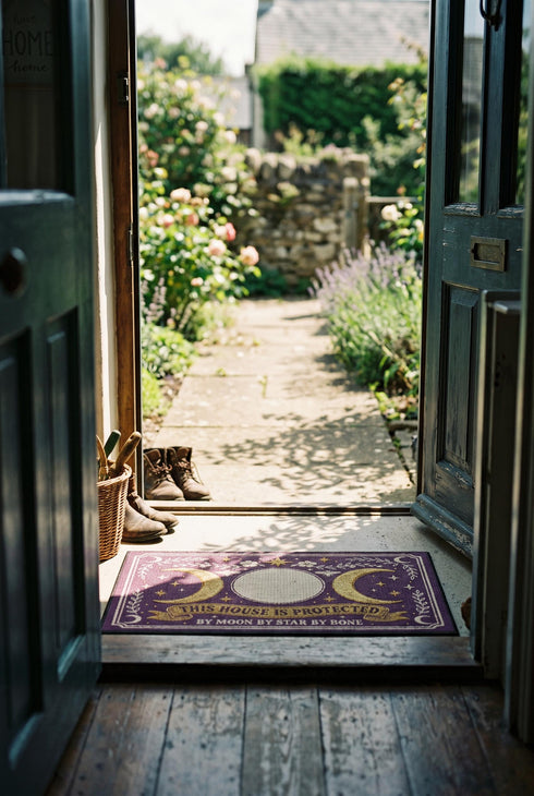 A rectangular doormat featuring a printed faux embroidery design on a deep plum purple background. The symmetrical artwork centers on a large cream full moon flanked by two golden crescent moons, creating a Triple Moon symbol. Above the moons are gold stars, cream flowers, and two small bees. Below, a golden scroll banner reads "THIS HOUSE IS PROTECTED" with "BY MOON BY STAR BY BONE" underneath. The mat is framed by cream leafy vines and finished with a black fabric border trim.