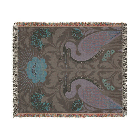 Jacquard woven cotton throw blanket, woven rather than printed, shown flat. Two mirrored great blue herons flank a central radial sunburst, surrounded by botanical blooms and scrollwork. The charcoal black ground carries antique gold ochre sunbeams, blush pink blossoms, deep forest green leaves, muted rust berry clusters, and cream toned herons. A lush multi-colored fringe of black, ochre, and sage threads edges the blanket, and the reverse shows an inverted charcoal grey version of the same design.