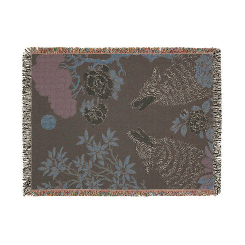 A high-contrast jacquard woven blanket featuring two wolves howling toward a central moon. The design is rendered in charcoal, sage green, and cream threads, showcasing intricate floral motifs like peonies and leafy trees. The textile has a visible woven texture with a multicolored fringe border. This Grimoire and Goblet piece displays a symmetrical, nature-inspired scene that functions as both a heavy throw and a wall tapestry, embodying a dark, botanical aesthetic.