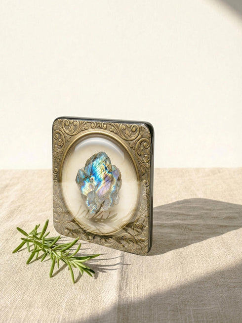 Grimoire and Goblet set of four square glossy coasters featuring high-resolution printed illustrations of raw crystals: purple amethyst, pink rose quartz, iridescent labradorite, and banded agate. Each gemstone is depicted inside a printed faux antique brass baroque frame with intricate scrollwork, set against a cream parchment background. The design is a flat 2D trompe l'œil print on hardboard, creating a realistic 3D specimen box illusion.
