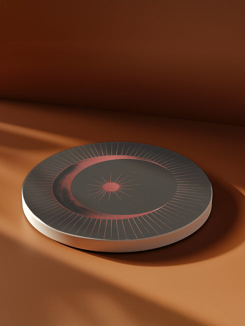 A round ceramic plate with a sleek design featuring a crescent moon and radiating lines in a muted red and black color scheme. The plate is set against a warm brown background with soft lighting, highlighting the celestial and minimalist aesthetic of the piece.