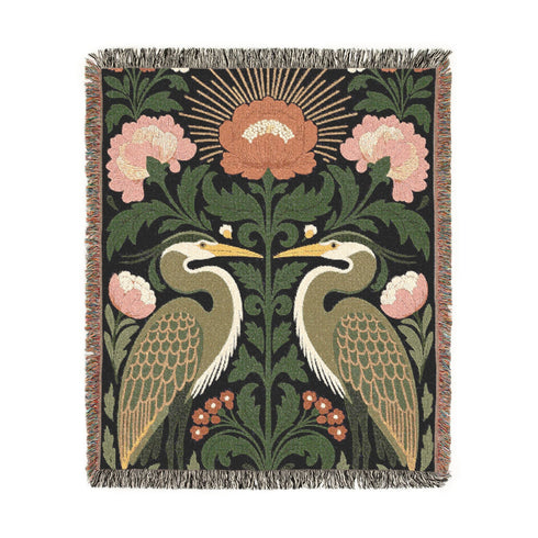 Jacquard woven cotton throw blanket, woven rather than printed, shown flat. Two mirrored great blue herons flank a central radial sunburst, surrounded by botanical blooms and scrollwork. The charcoal black ground carries antique gold ochre sunbeams, blush pink blossoms, deep forest green leaves, muted rust berry clusters, and cream toned herons. A lush multi-colored fringe of black, ochre, and sage threads edges the blanket, and the reverse shows an inverted charcoal grey version of the same design.