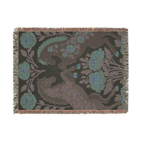 A woven cotton throw blanket by Grimoire and Goblet featuring two rearing horses facing each other in a symmetrical folk art design. One horse is charcoal black with rust floral detailing; the other is navy blue with teal floral detailing. They are framed by large rust-colored peonies and green leafy vines on a cream background. The blanket features a multicolor fringe edge. Perfect for dark academia and cottagecore aesthetics.