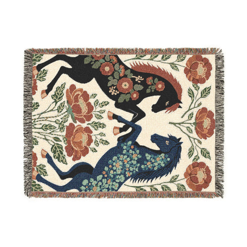A woven cotton throw blanket by Grimoire and Goblet featuring two rearing horses facing each other in a symmetrical folk art design. One horse is charcoal black with rust floral detailing; the other is navy blue with teal floral detailing. They are framed by large rust-colored peonies and green leafy vines on a cream background. The blanket features a multicolor fringe edge. Perfect for dark academia and cottagecore aesthetics.
