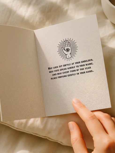 Grimoire and Goblet presents a 5x7 inch pearlescent greeting card. The design features a black linocut-style illustration of an open palm with an eye, surrounded by rays, keys, coins, and stars. The cover reads Luck Fate Fortune and May The Coming Year Favor Your Hand. The interior features a smaller hand icon and a printed blessing. The paper has a luxury silvery shimmer finish.