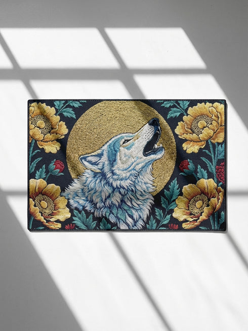Grimoire and Goblet rectangular doormat featuring a faux embroidery style printed design of a howling wolf with blue and ivory fur, centered in front of a textured antique gold circular moon disc, surrounded by large golden yellow flowers, teal leaves, and small red berry accents on a dark charcoal background. Entryway rug with black trim, non slip backing, and botanical nature inspired mystical design.
