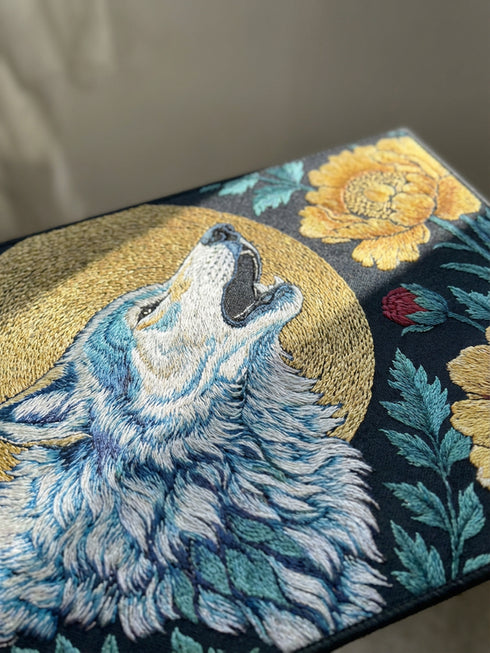 Grimoire and Goblet rectangular doormat featuring a faux embroidery style printed design of a howling wolf with blue and ivory fur, centered in front of a textured antique gold circular moon disc, surrounded by large golden yellow flowers, teal leaves, and small red berry accents on a dark charcoal background. Entryway rug with black trim, non slip backing, and botanical nature inspired mystical design.