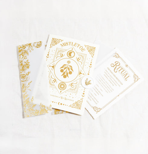 A Grimoire & Goblet luxury Mistletoe ritual card set. The 5x7 cream greeting card features gold celestial designs framing “Mistletoe” and “Love, Protection & New Beginnings.” Shown with a vellum wrap printed in gold florals, a white wax seal with mistletoe, and a premium envelope. Includes a 4x6 ritual insert on handmade deckled paper titled “Ritual for Peace and Protection.” A spiritual Yule stationery set designed as a luxurious unboxing experience.