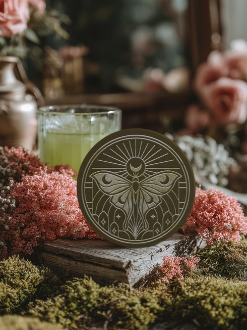 A round, dark brown ceramic drink coaster with a non-slip cork back. The top surface features an intricate, engraved-style celestial moth design in a light beige or off-white color. The luna moth is centered under a crescent moon and a radiant sunburst. The design is symmetrical and includes stars and geometric line art, evoking a gothic, witchy, and spiritual aesthetic. The coaster has a matte finish and is designed as a piece of functional art.