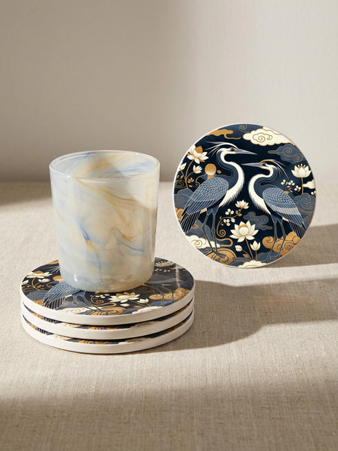 A round 4-inch drink coaster by Grimoire and Goblet. The coaster features a highly reflective glossy top surface displaying two Great Blue Herons, lotus flowers, and stylized clouds in metallic antique gold and slate blue against a solid, deep navy blue background. The edges reveal a natural brown, porous cork backing underneath. The intricate design resembles vintage Chinoiserie and botanical natural history art, perfectly suited for protecting tables from beverages.