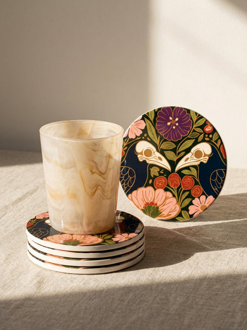 A square drink coaster with a high-gloss finish and natural cork backing. The centralized, symmetrical design features two detailed, cream-colored avian bird skulls facing inward. They are framed by lush botanical flora, including a large purple flower, blush pink blooms, rust-red roses, and green ferns, all set against a deep navy blue background with gold linework accents. Functional gothic, whimsigoth, and dark academia drinkware designed by the Grimoire and Goblet brand.